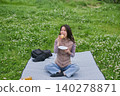 A woman eats a sandwich on a picnic blanket spread out on a grassy field with fresh green leaves. 140278871