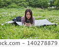 A woman is looking at her smartphone while sitting on a sheet spread out on a grassy field with fresh green leaves. 140278879