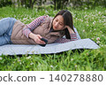 A woman is looking at her smartphone while sitting on a sheet spread out on a grassy field with fresh green leaves. 140278880