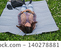 A woman is looking at her smartphone while sitting on a picnic blanket in a grassy area with fresh green leaves. 140278881