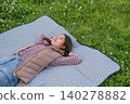 A woman lies down on a picnic blanket spread out in a grassy area with fresh green leaves. 140278882