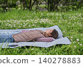 A woman lies down on a picnic blanket spread out in a grassy area with fresh green leaves. 140278883