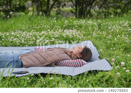 A woman lies down on a picnic blanket spread out in a grassy area with fresh green leaves. A woman lies down on a picnic blanket spread out in a grassy area with fresh green leaves. 140278883