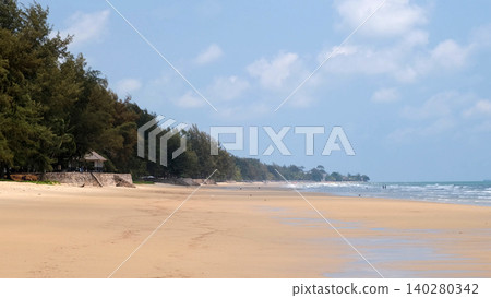 Long and wide sandy beach with calm blue sea and lush tropical pine trees along the shoreline under blue sky. Tropical beach concept Long and wide sandy beach with calm blue sea and lush tropical pine trees along the shoreline under blue sky. Tropical beach concept 140280342