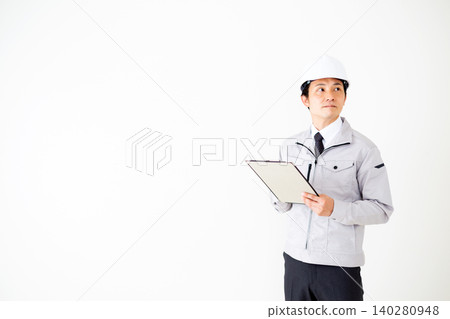 A middle-aged businessman in gray work clothes and a helmet, holding a binder of documents, against a white background. A middle-aged businessman in gray work clothes and a helmet, holding a binder of documents, against a white background. 140280948