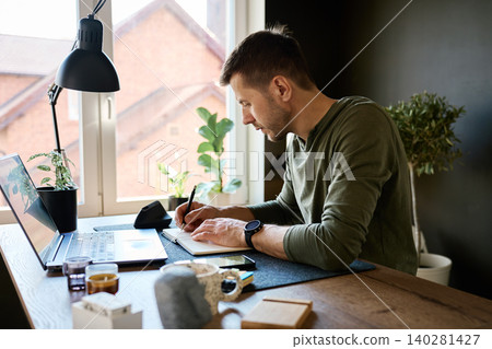 Man writing in notebook at home workspace with laptop by window Man writing in notebook at home workspace with laptop by window 140281427