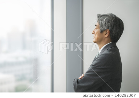 一位六十多歲的商人，雙手交叉抱在胸前，望著窗外（側面照） 140282307