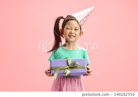 Asian ethnicity little girl appearance with party hat and festive clothes hugging her birthday present, that her dream had finally come true beaming with joy standing over pink isolated. Asian ethnicity little girl appearance with party hat and festive clothes hugging her birthday present, that her dream had finally come true beaming with joy standing over pink isolated. 140282550