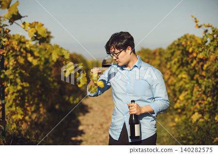 Man Tasting Red Wine from Glass Standing in Sunlit Vineyard 140282575