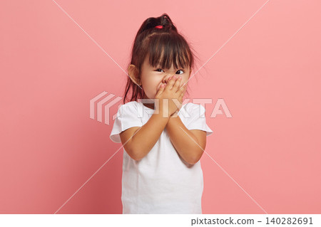 Little Asian girl covers her mouth with her hand with her eyes wide open in shame, said something superfluous and inappropriate, expresses embarrassment and shyness while standing on a pink isolated. Little Asian girl covers her mouth with her hand with her eyes wide open in shame, said something superfluous and inappropriate, expresses embarrassment and shyness while standing on a pink isolated. 140282691