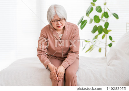 A senior woman in pajamas sits on her bed after waking up, complaining of knee pain (knee, knee pain). A senior woman in pajamas sits on her bed after waking up, complaining of knee pain (knee, knee pain). 140282955
