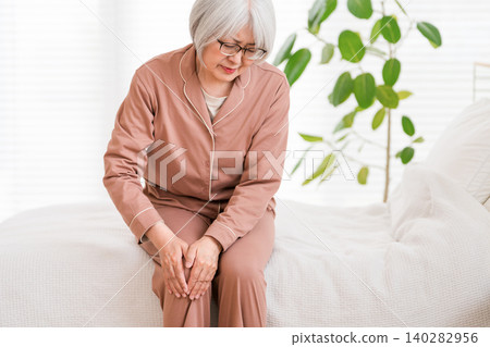 A senior woman in pajamas sits on her bed after waking up, complaining of knee pain (knee, knee pain). A senior woman in pajamas sits on her bed after waking up, complaining of knee pain (knee, knee pain). 140282956