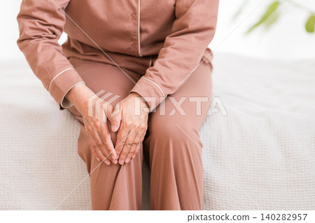 A senior woman in pajamas sits on her bed after waking up, complaining of knee pain (knee, knee pain). 140282957