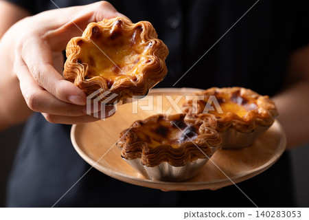 Egg tarts filled with creamy custard and caramelized tops on a wooden plate holding by hand Egg tarts filled with creamy custard and caramelized tops on a wooden plate holding by hand 140283053