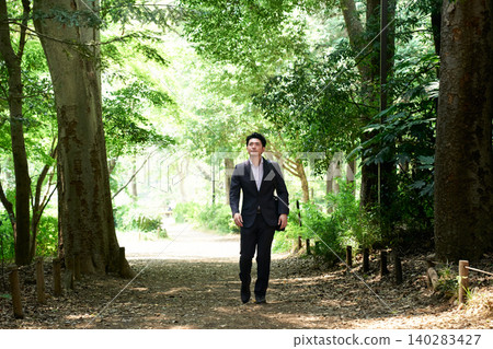 A Japanese man in his 30s, on his outside sales job, is strolling along a shady path in a lush park, looking up at the sky with a smile on his face. A Japanese man in his 30s, on his outside sales job, is strolling along a shady path in a lush park, looking up at the sky with a smile on his face. 140283427