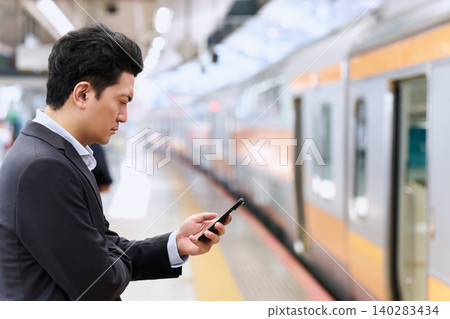 一名三十多歲的日本男子，身穿西裝，在通勤途中，於火車站台上專注地操作他的智慧型手機。 140283434