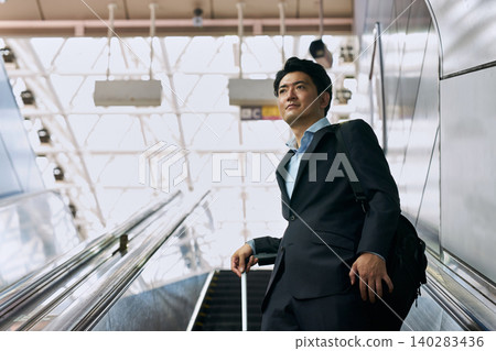 A Japanese businessman in his 30s, wearing a suit and carrying a carry-on bag, is seen on an escalator during a business trip. 140283436
