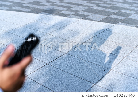 In an urban outdoor setting, the shadow of an office worker holding a smartphone is projected onto the cobblestone ground, revealing the faceless hands. In an urban outdoor setting, the shadow of an office worker holding a smartphone is projected onto the cobblestone ground, revealing the faceless hands. 140283440