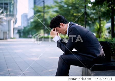 A Japanese office worker in his 20s sits on an outdoor bench in the city, his head in his hands, troubled by work-related stress. A Japanese office worker in his 20s sits on an outdoor bench in the city, his head in his hands, troubled by work-related stress. 140283441