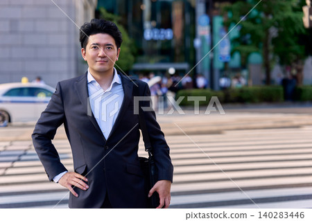 一名三十多歲的日本男子，公司職員，站在城市的斑馬線上，雙手叉腰，面帶微笑，直視著鏡頭。 140283446