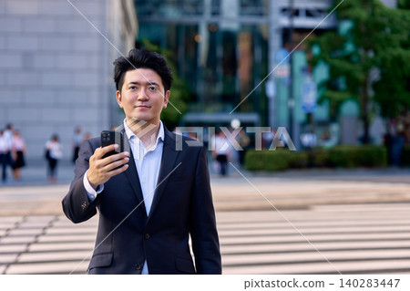 一名三十多歲的日本男子,公司職員,傍晚時分站在城市辦公區的十字路口,手裡拿著他的智慧型手機。 一名三十多歲的日本男子,公司職員,傍晚時分站在城市辦公區的十字路口,手裡拿著他的智慧型手機。 140283447