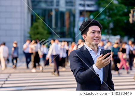 一位三十多歲的日本商人,在尖峰時段的辦公區一邊操作智慧型手機一邊微笑。 一位三十多歲的日本商人,在尖峰時段的辦公區一邊操作智慧型手機一邊微笑。 140283449