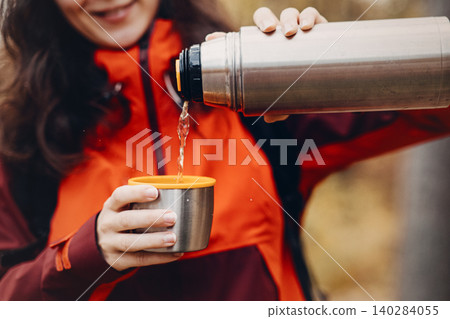 Smiling woman pours hot beverage from metal thermos into cup outdoors. seasonal forest recreation, functional camping outfit, cold weather trekking, stylish outdoor gear. Smiling woman pours hot beverage from metal thermos into cup outdoors. seasonal forest recreation, functional camping outfit, cold weather trekking, stylish outdoor gear. 140284055
