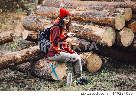Woman sits on log in forest and looks at smartphone screen. Smartphone usage in forest, mobile roaming for tourists, digital navigation tools. Woman sits on log in forest and looks at smartphone screen. Smartphone usage in forest, mobile roaming for tourists, digital navigation tools. 140284056