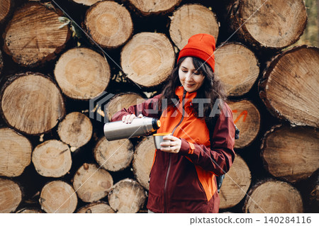 Smiling woman pours drink from metal thermos into cup while standing in front of stacked logs in forest. Independent female traveler, solo outdoor living, Autonomous nature exploration. 140284116