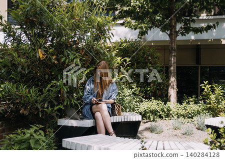 Woman sits on bench in green courtyard writing in notebook next to laptop computer. Connected work lifestyle, mobile technology productivity, healthy workspace choice. Woman sits on bench in green courtyard writing in notebook next to laptop computer. Connected work lifestyle, mobile technology productivity, healthy workspace choice. 140284128