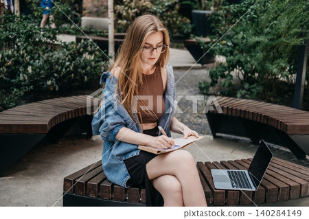 Woman writes in notepad near laptop while sitting on wooden public bench outdoors. Digital nomad life, laptop on bench, modern work solutions, Tech-enabled lifestyle. 140284149