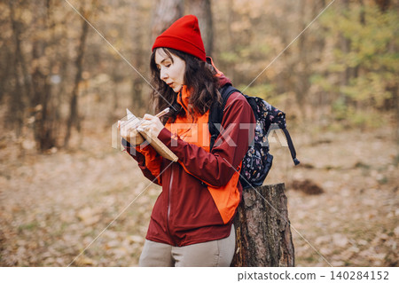 Woman stands in forest while writing in notebook and leaning on tree stump. Nature journaling therapy, mindfulness forest walking, mental health recovery. Woman stands in forest while writing in notebook and leaning on tree stump. Nature journaling therapy, mindfulness forest walking, mental health recovery. 140284152