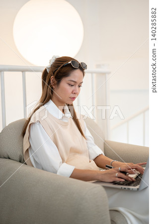 Asian woman holding pen as typing on laptop over legs while sitting on sofa chair in cafe or at home Asian woman holding pen as typing on laptop over legs while sitting on sofa chair in cafe or at home 140285882