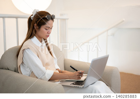 Asian woman holding pen writing in book over laptop on legs while sitting on sofa in cafe or at home 140285883