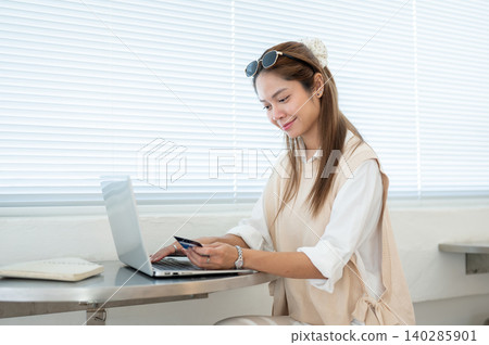 Asian woman holding looking at credit card over laptop while sitting at table in cafe or home office Asian woman holding looking at credit card over laptop while sitting at table in cafe or home office 140285901