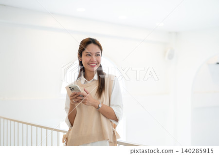 Smiling asian woman looking away from phone as standing leaning against railing in cafe or at home. 140285915