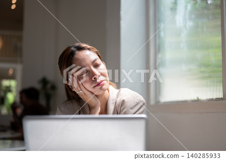 Asian woman closing eyes resting head on hand under sunlight sitting with laptop beside cafe window. Asian woman closing eyes resting head on hand under sunlight sitting with laptop beside cafe window. 140285933