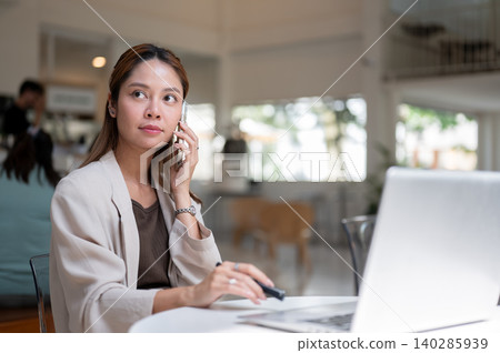 Pretty asian woman holding pen making a phone call in front of laptop while sitting at table in cafe Pretty asian woman holding pen making a phone call in front of laptop while sitting at table in cafe 140285939