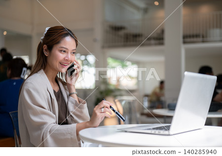 Pretty asian woman holding pen talking on phone call in front of laptop as sitting at table in cafe. Pretty asian woman holding pen talking on phone call in front of laptop as sitting at table in cafe. 140285940