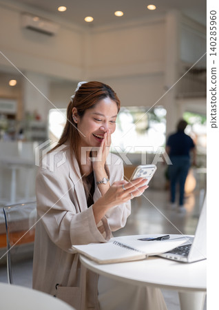 Happy asian woman looking at phone covering smiling mouth over laptop while sitting at table in cafe Happy asian woman looking at phone covering smiling mouth over laptop while sitting at table in cafe 140285960