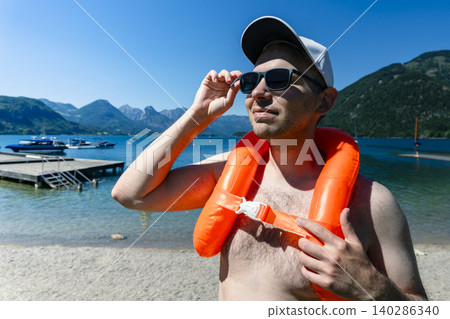 Man wearing an orange inflatable life vest and sunglasses on a sunny beach, highlighting water safety during outdoor recreation Man wearing an orange inflatable life vest and sunglasses on a sunny beach, highlighting water safety during outdoor recreation 140286340