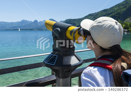 Woman looking through a coin operated telescope at mountains and lake Wolfgangsee in Austria, emphasizing outdoor observation 140286372