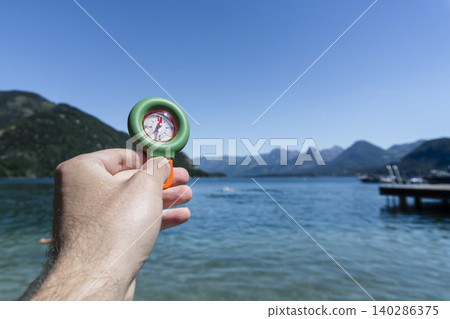 Hand holding a portable compass over a lake with mountains in the background, suitable for travel use and practical field tasks Hand holding a portable compass over a lake with mountains in the background, suitable for travel use and practical field tasks 140286375
