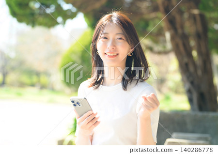 一位日本女子在公園裡開心地拿著她的智慧型手機 一位日本女子在公園裡開心地拿著她的智慧型手機 140286778