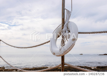 White lifebuoy hanging on a wooden post and rope fence beside the Mediterranean Sea in Paphos, Cyprus. White lifebuoy hanging on a wooden post and rope fence beside the Mediterranean Sea in Paphos, Cyprus. 140287236