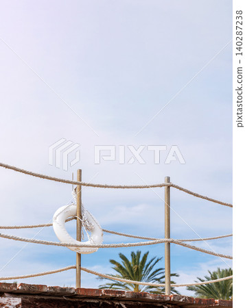 White lifebuoy on a rope fence by Mediterranean seaside in Paphos, Cyprus. Minimal resort setting with cloudy sky, palm tree. White lifebuoy on a rope fence by Mediterranean seaside in Paphos, Cyprus. Minimal resort setting with cloudy sky, palm tree. 140287238