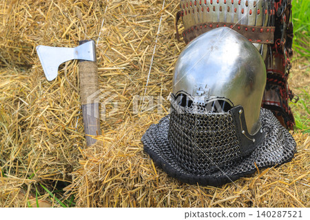 metallic medieval helmet chainmail hatchet axe placed on a rustic hay bale evoking a historical combat scene metallic medieval helmet chainmail hatchet axe placed on a rustic hay bale evoking a historical combat scene 140287521