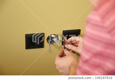 Close-up of electrician hands stripping electrical wires for socket installation. Electrician using utility knife blade to repair power outlet in a yellow wall, domestic home renovation concept Close-up of electrician hands stripping electrical wires for socket installation. Electrician using utility knife blade to repair power outlet in a yellow wall, domestic home renovation concept 140287918