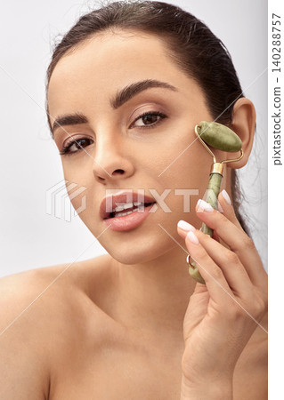 Facial jade roller. Young woman massages her sensuality face using a green jade massager roller for elastic perfect skin Facial jade roller. Young woman massages her sensuality face using a green jade massager roller for elastic perfect skin 140288757
