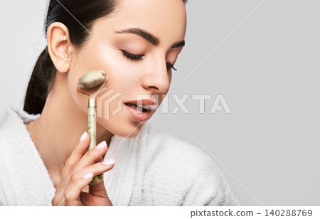 Facial jade roller. Young woman massages her face using a green jade roller for elastic perfect skin. Isolated on grey 140288769